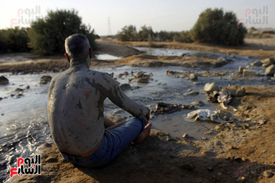   رجل  يجلس علي طرف بركة المياه الكبريتية  ينتظر  ان  يجف جسمه بعدما دهنه  طين  من البركة  