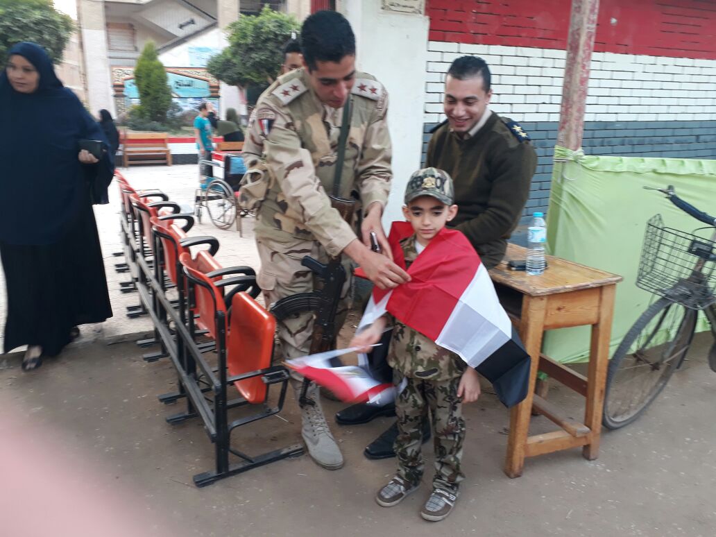 أحد رجال الجيش المكلفين بالتأمين يساعد الطفل بإرتداء العلم
