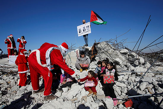متظاهر يرتدى زى بابا نويل يوزع هدايا على الأطفال الجالسين فوق أنقاض منزل