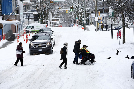 المشاة يساعدون رجلاً يستخدم كرسيًا متحركًا في عبور الشارع بعد عاصفة ثلجية في وسط مدينة فانكوفر