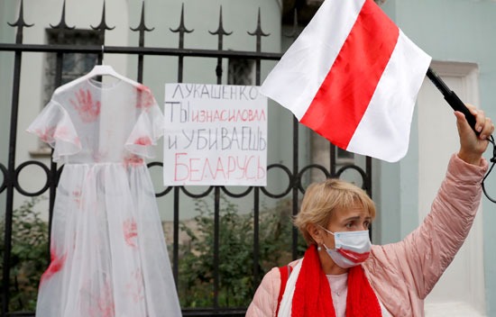 امرأة تحمل علم بيلاروسيا وفي الخلفية فستان أبيض عليه كفوف حمراء